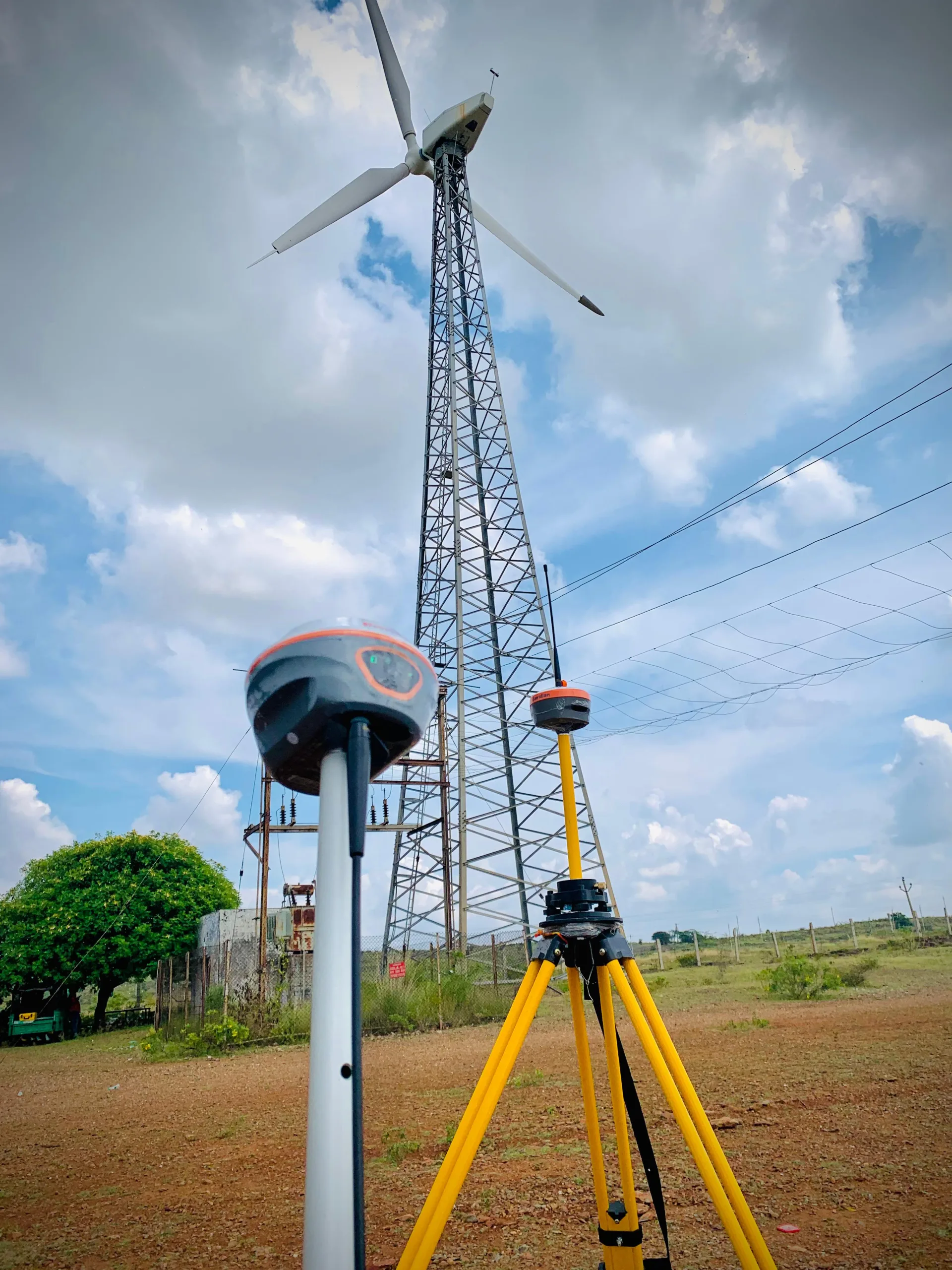 GNSS rover and tripod set up beside a wind turbine and transmission tower